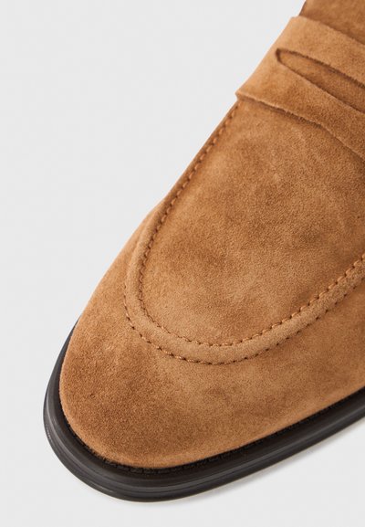 Smeđi mokasinski cipele od brušene kože s zaobljenim vrhom, prošivenim detaljima i teksturiranim gornjim dijelom. Imaju crni gumeni potplat za bolji grip i trajnost.