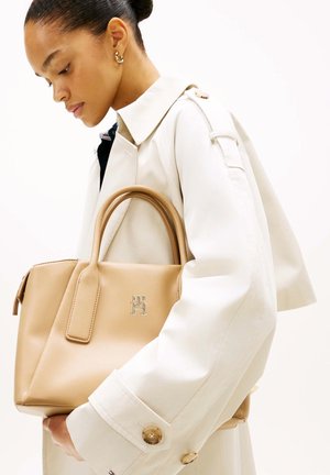 Vrouw met haar in een knot houdt een beige leren handtas vast, draagt een lichtbeige trenchcoat en gouden oorbellen in de vorm van hoepels tegen een witte achtergrond.