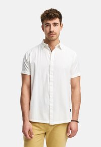 Camisa blanca de manga corta con un patrón texturizado, frente abotonado y sutiles acentos de color en el collar. Combinada con pantalones amarillos.