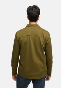 Camicia a maniche lunghe verde oliva con colletto, tessuto strutturato e dettaglio di cucitura sulla schiena. Indossata con jeans blu scuro.