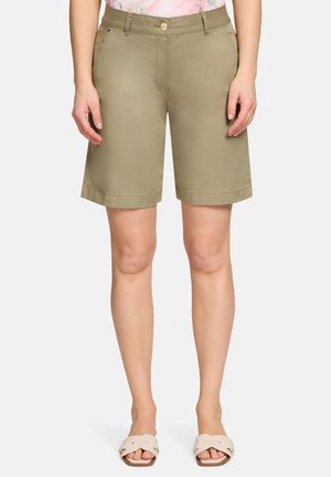 Vrouw draagt lichtbruine shorts tot op de knie met zijzakken en een knoopsluiting, gecombineerd met beige instapsandalen.