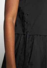 Närbild av en persons arm bredvid en svart topp med flätad snörningsdetalj på sidan och en texturerad svart kjol.