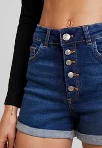 Jeansshorts i mörk tvätt med hög midja, med fem metalldragknappar och beige sömnad. Uppvikt fåll i botten.