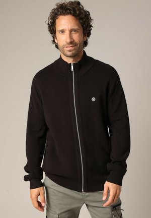 Sweat à zipper noir avec un col montant, fabriqué en matière tricot douce. Dispose d'un logo rond sur la poitrine gauche et de poignets ajustés.
