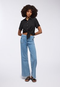 Camicia nera a maniche corte con bottoni e nodo frontale, abbinata a jeans in denim azzurri chiari a gamba larga con due tasche posteriori e cuciture piatte.