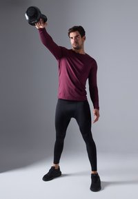 Bordo ilgo rankovių marškiniai, juodos aptemptos legging'ės ir juodi sportiniai bateliai. Asmuo laiko 10 kg svorį, turėdamas sportinę poziciją prieš neutralų foną.