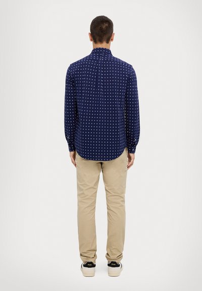 Vyras stovi nuo nugaros, vilkintis tamsiai mėlyną raštuotą ilgarankovį marškinį, smėlio spalvos kelnes ir baltus "Polo" sportbačius, prieš paprastą foną.