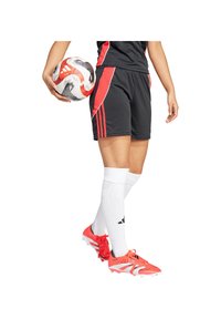 Giocatore di calcio con pantaloni corti neri e rossi, calze bianche fin sopra il ginocchio e scarpe arancioni tiene in mano un pallone da calcio bianco e rosso.