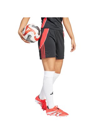 Ein Fußballspieler in schwarzen und roten Shorts, weißen Kniestrümpfen und orangenen Fußballschuhen hält einen weißen und roten Fußball an seiner Seite.