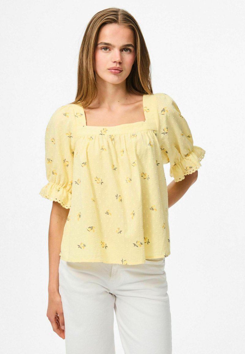 Blusa gialla con maniche a palloncino e scollatura quadrata, tessuto testurizzato, piccolo motivo floreale e bordo scallopato alle maniche. Abbinata a pantaloni bianchi.