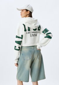 Haut à capuche blanc court avec lettrage vert "CALIFORNIA LOS ANGELES 1996", associé à un short en denim léger bleu délavé. Boucle sur la taille.