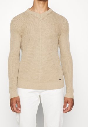 Homme portant un pull en tricot beige à manches longues avec un col en V subtil, associé à un pantalon blanc, debout devant un fond uni.