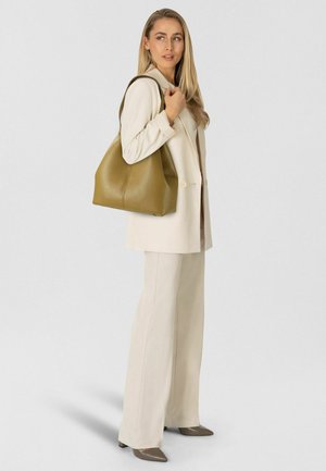 Sac cabas en cuir vert à finition texturée, doté d'une large ouverture et de courbes douces, tenu par une personne portant un costume beige clair.