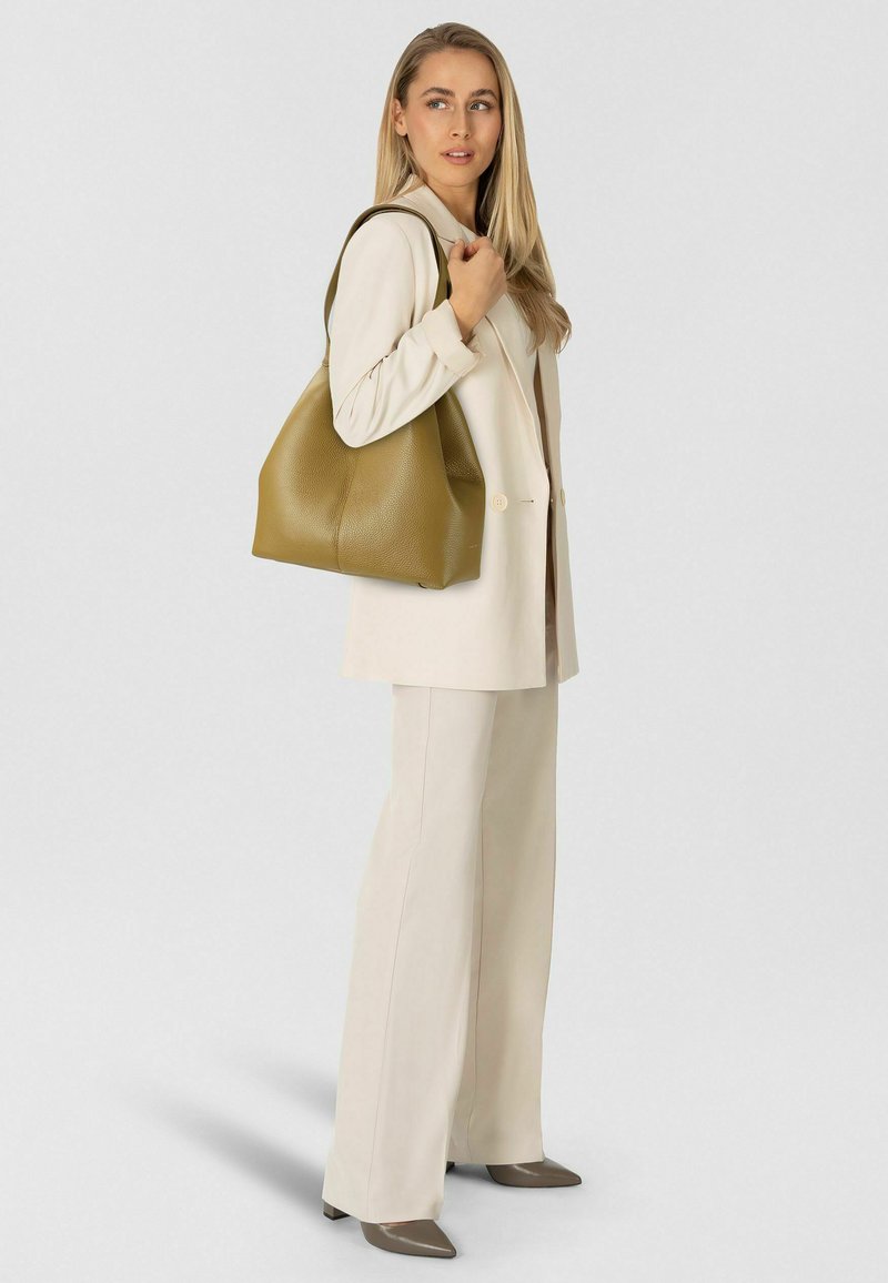 Borsa a mano in pelle verde con finitura strutturata, caratterizzata da un'apertura ampia e curve morbide, tenuta da una persona con un abito beige chiaro.