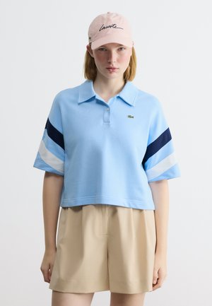 Γυναίκα που φοράει ένα γαλάζιο κοντό πόλο με ναυτικά και λευκά ριγέ, μπεζ σόρτς και ρόζ καπέλο με μαύρο κείμενο στο μπροστινό μέρος.