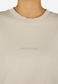 Šviesiai smėlinis džemperis su groteliniu apykaklės kraštu ir tekstūruota medžiaga. Priekyje yra subtilus, įspaustas logotipas.