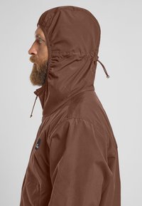 Bruine hooded jas met een trekkoordcapuchon, met een gladde textuur, zijzakken en een logo-opdruk op de borst, gepresenteerd op een grijze achtergrond.