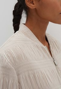 Helle beigetfarbene Jacke mit einer zerknitterten Textur, ausgestattet mit einem Stehkragen und silbernem Reißverschluss. Der Stoff weist horizontale Falten für zusätzlichen Detailreichtum auf.