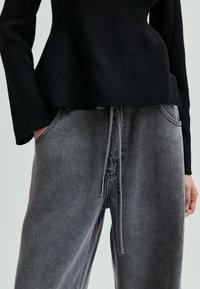 En person klädd i en svart långärmad ribbad topp och lösa grå denimbyxor med dragsko, med händerna vilande i framfickorna.