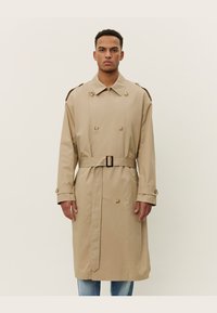 Trench coat beige en tela suave con diseño de doble botonadura, grandes botones, epaulettes en los hombros y cinturón en la cintura.