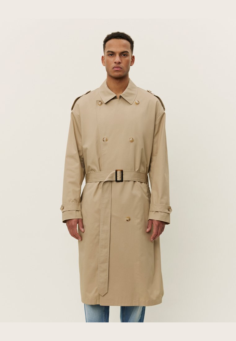 Trench coat beige en tela suave con diseño de doble botonadura, grandes botones, epaulettes en los hombros y cinturón en la cintura.