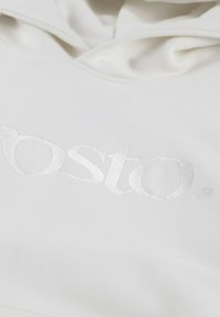 Biała bluza z kapturem, charakteryzująca się miękką teksturą oraz wyraźnym, odblaskowym wytłoczonym logo na środku. Projekt jest prosty i elegancki.