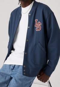 Chaqueta de estilo varsity en azul marino con botones a presión, logo bordado "LS" en burdeos, con cuello redondo y bolsillos laterales.