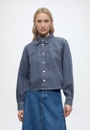 Γυναίκα με ανοιχτόχρωμα μαλλιά που φοράει γκρι μπουφάν από τζιν με κουμπιά και μπλε φούστα από τζιν, στέκεται μπροστά σε απλό φόντο.