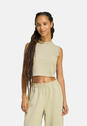 Junge Frau mit langem geflochtenem Haar, die ein ärmelloses, bauchfreies Oberteil und lockere Hose mit elastischem Bund in hellem Beigeton trägt, stehend und lächelnd.