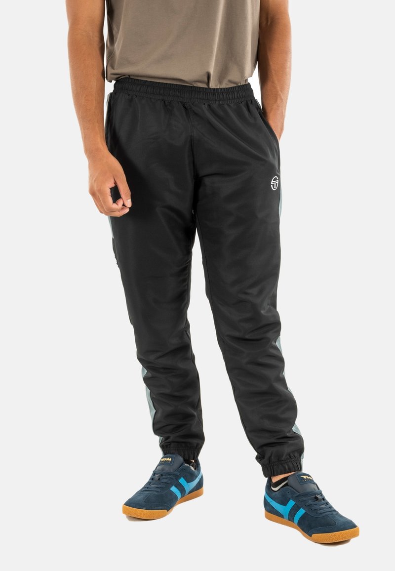 Pantaloni da jogging neri con vita elastica, gambe affusolate e pannelli laterali in turchese. Presentano un logo sulla coscia sinistra. Indossati con sneakers blu.