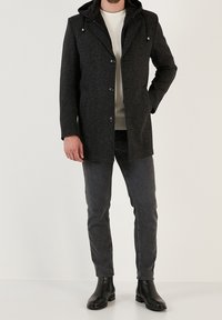 Abrigo negro de tela texturizada con capucha desmontable. Cuenta con botones frontales, bolsillos laterales y un corte entallado. Llevado con jeans grises y botas negras.