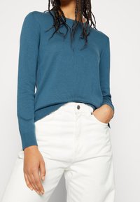 Türkisfarbener Strickpullover mit langen Ärmeln und strukturiertem Stoff, kombiniert mit hoch taillierten weißen Jeans mit Knopfverschluss und Vordertaschen.