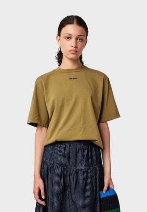 T-shirt in cotone verde oliva con collo a giro e maniche corte, con un piccolo logo blu sul petto. Modello abbinato a una gonna a righe.