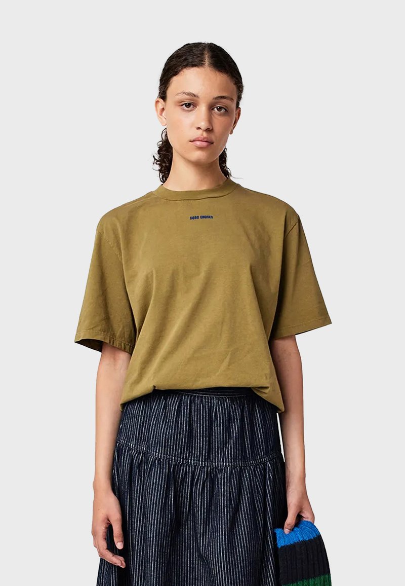 Kvinna med lockigt hår bär en olivgrön oversized t-shirt och en högmidjad mörkblå veckad kjol, håller i en blå, svart och grön artikel.