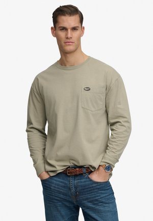 Mężczyzna ubrany w beżową koszulę z długim rękawem i kieszenią na piersi, niebieskie dżinsy, pleciony brązowy pasek oraz zegarek na rękę, stojący z rękami w kieszeniach.