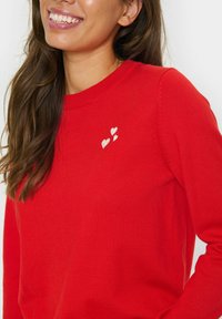 Röd stickad tröja med rund halsringning och tre broderade hjärtan i vitt på vänster sida. Mjuk textur och figursydd design.
