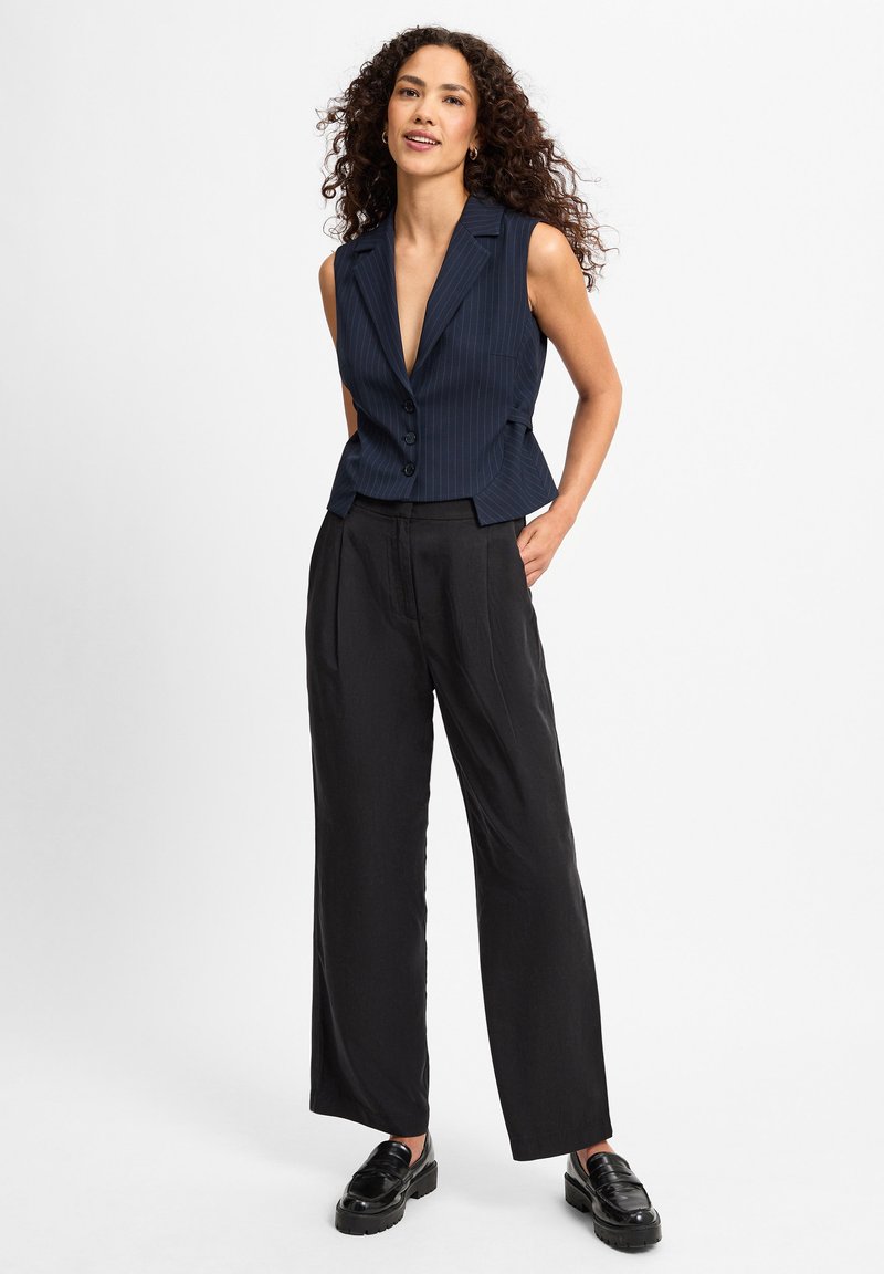 Marineblau gestreifter ärmelloser Blazer mit tiefem V-Ausschnitt und vier Knöpfen, kombiniert mit schwarzen weit geschnittenen Hosen und glänzenden schwarzen Loafers.