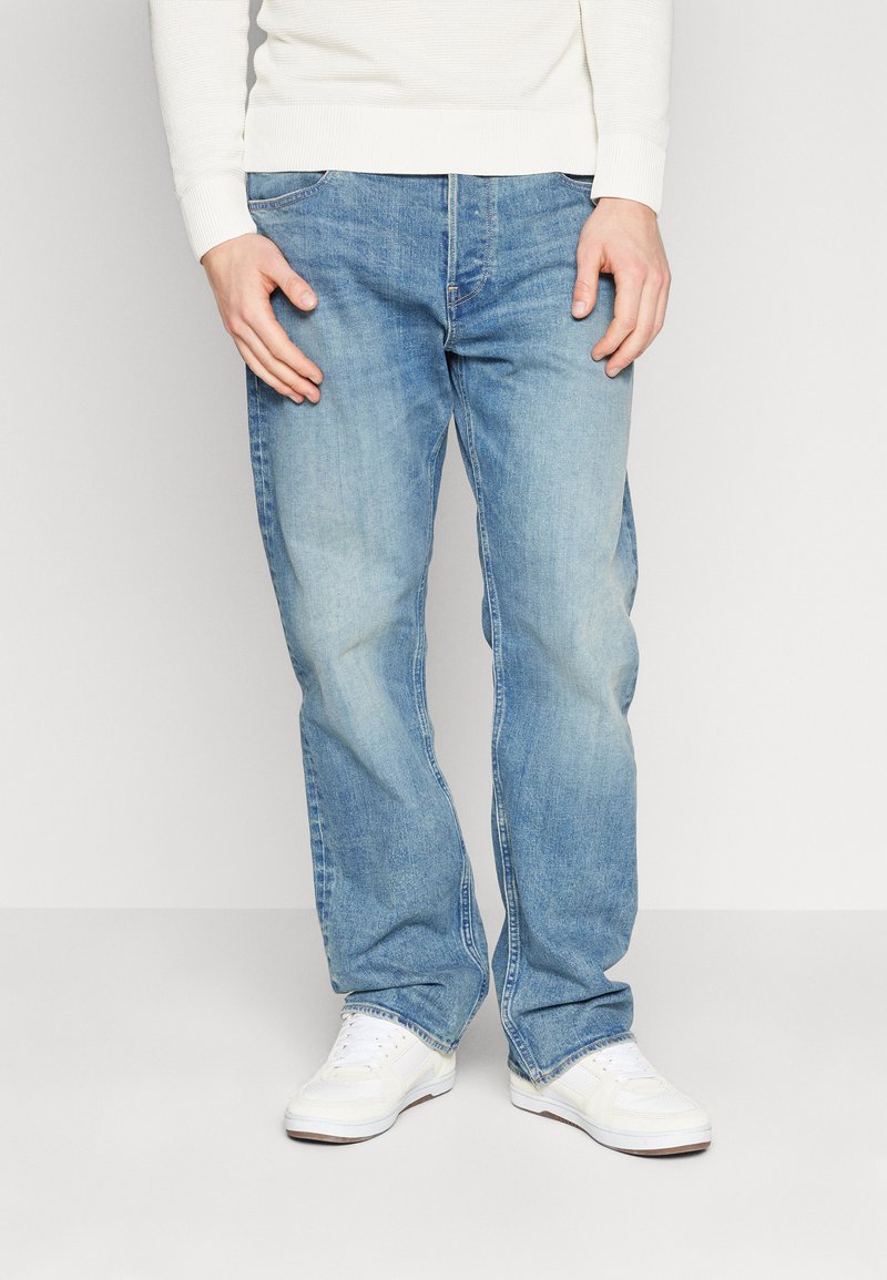 Jeans de denim azul con un corte recto, lavado claro, desvanecimiento sutil y diseño clásico de cinco bolsillos, combinados con zapatillas blancas.