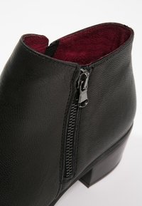 Bottine en cuir noir avec fermeture éclair latérale et doublure en tissu bordeaux, dotée d'un petit talon bloc et d'une surface texturée.