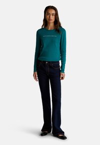 Tee blaugrüne Langarmshirt mit dem Aufdruck "United Colors of Benetton", kombiniert mit dunklen Schlagjeans und schwarzen Ballerinas. Einfaches Design, glatte Textur.