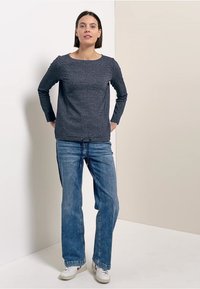 Frau steht mit hinter dem Rücken verschränkten Händen, trägt ein dunkel strukturiertes Langarm-Oberteil, blaue Jeans und weiße Turnschuhe vor einem neutralen Hintergrund.