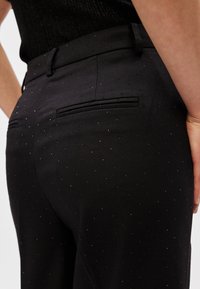 Melnas bikses ar gludu audumu ar smalku mirdzumu, rotātas ar maziem atstarojošiem akcentiem. Redzami divi aizmugurējie kabatas.