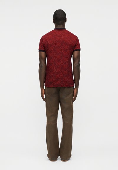 Άνδρας που στέκεται με την πλάτη προς την κάμερα, φοράει εφαρμοστό κόκκινο πουκάμισο με σχέδια και κοντά μανίκια και φαρδιά καφέ παντελόνια, μπροστά σε απλό φόντο.