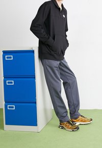 Felpa nera con zip e tasca frontale, pantaloni sportivi grigi e scarpe da ginnastica gialle e nere. Posizionati accanto a un armadietto blu.