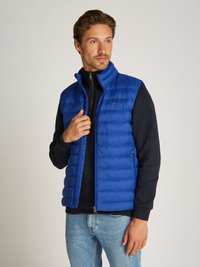 Blå puffer vest med quiltet tekstur, glidelås, og høy krage, brukt over en svart genser, sammen med lyse denimjeans.