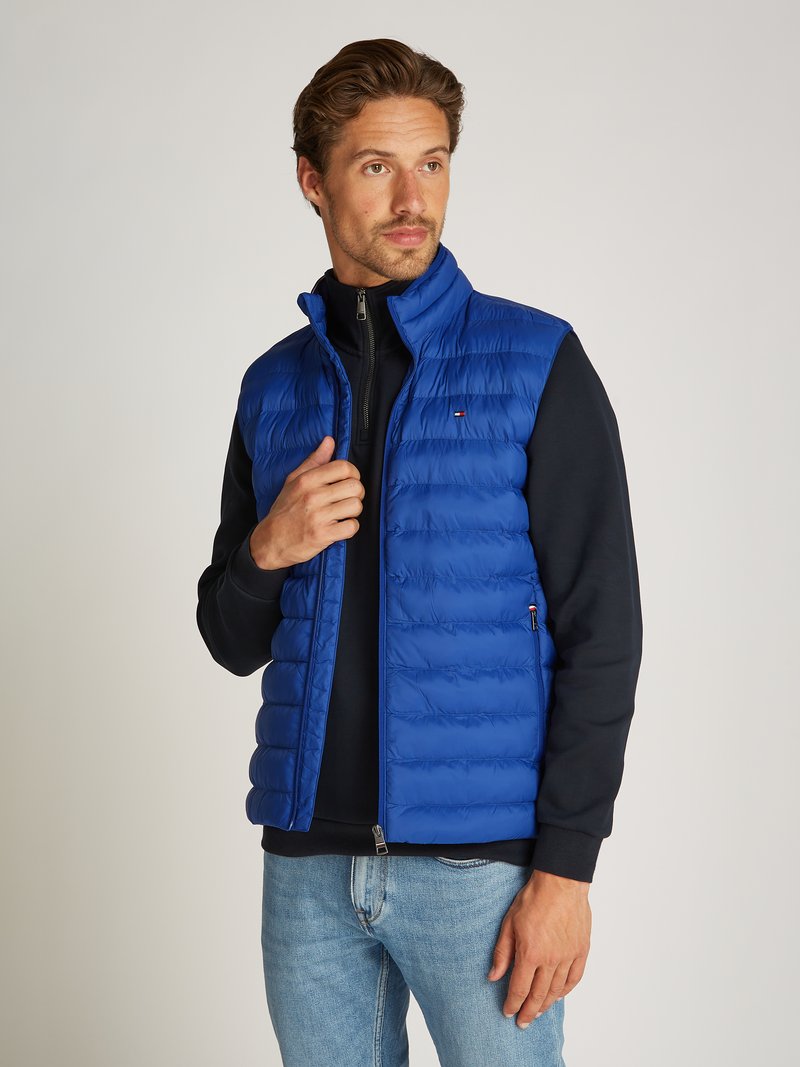 Blå puffer vest med quiltet tekstur, glidelås, og høy krage, brukt over en svart genser, sammen med lyse denimjeans.