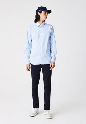 Camisa de botones azul claro con mangas largas, combinada con pantalones oscuros color marino y zapatillas blancas. Una gorra marina con un logo verde completa el aspecto.