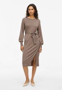 Robe marron à manches longues avec un design enveloppant et une fente latérale, dotée d'une taille nouée et d'une texture douce. Portez-la avec des talons beiges.