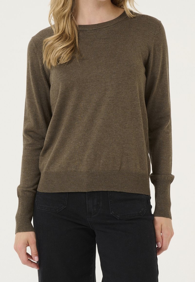 Femme portant un pull à manches longues vert olive et un jean noir, debout devant un fond clair uni.