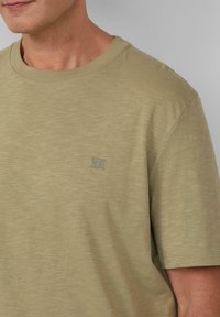 Jasnooliwkowa koszulka z miękkiego materiału, z okrągłym dekoltem i małym, wytłoczonym logo po lewej stronie klatki piersiowej.